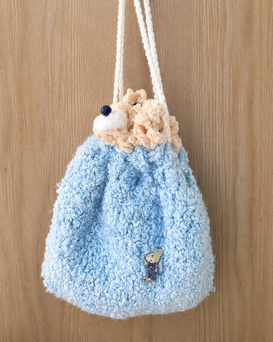 Styled product view of Madhu's handmade light blue yarn handbag with a peach-and-white animal face, cream handles, and a small mouse pin